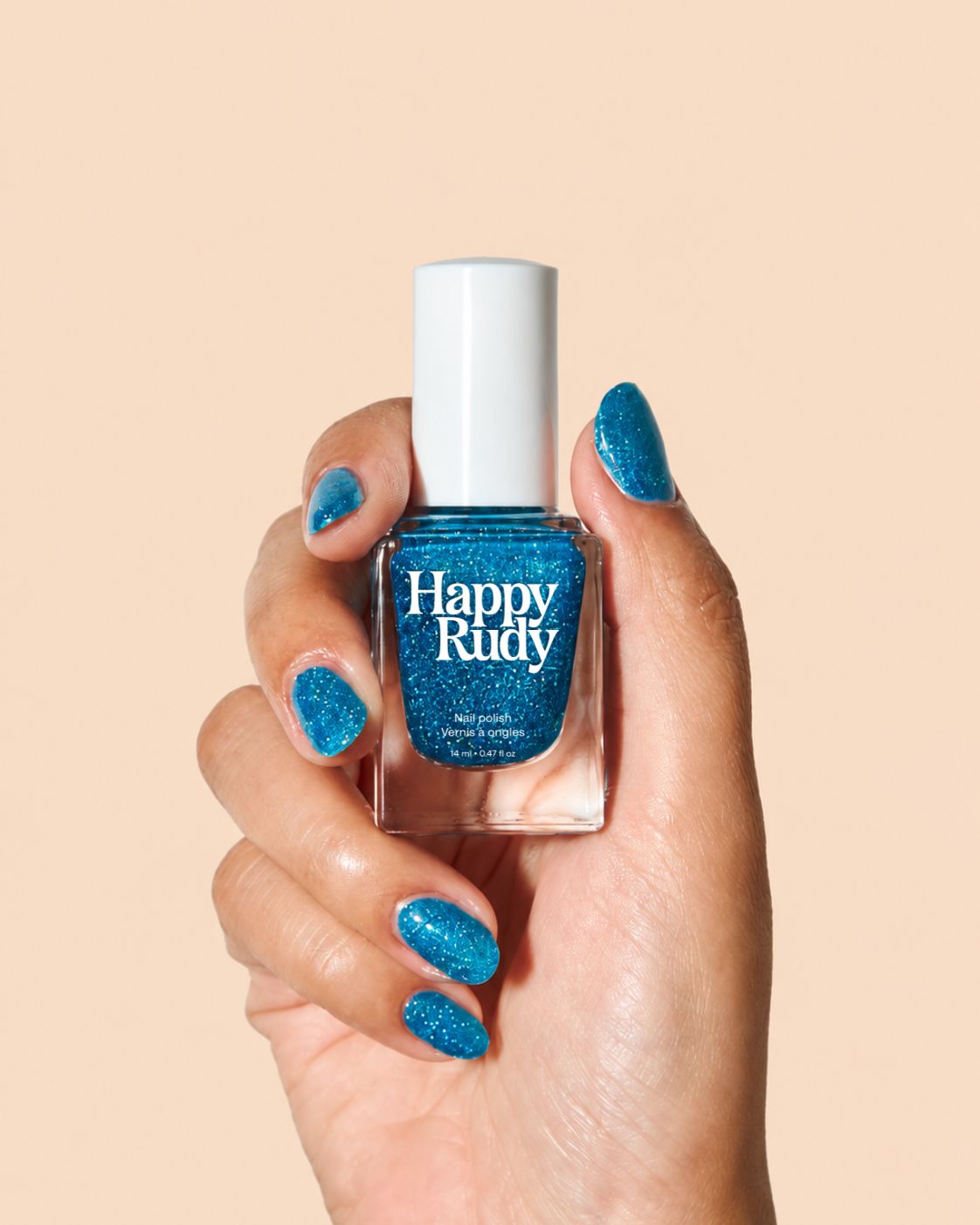 A hand holding a bottle of Happy Rudy glitter nail polish in a bright blue shade, with matching glittery blue nails painted on, against a solid beige background.