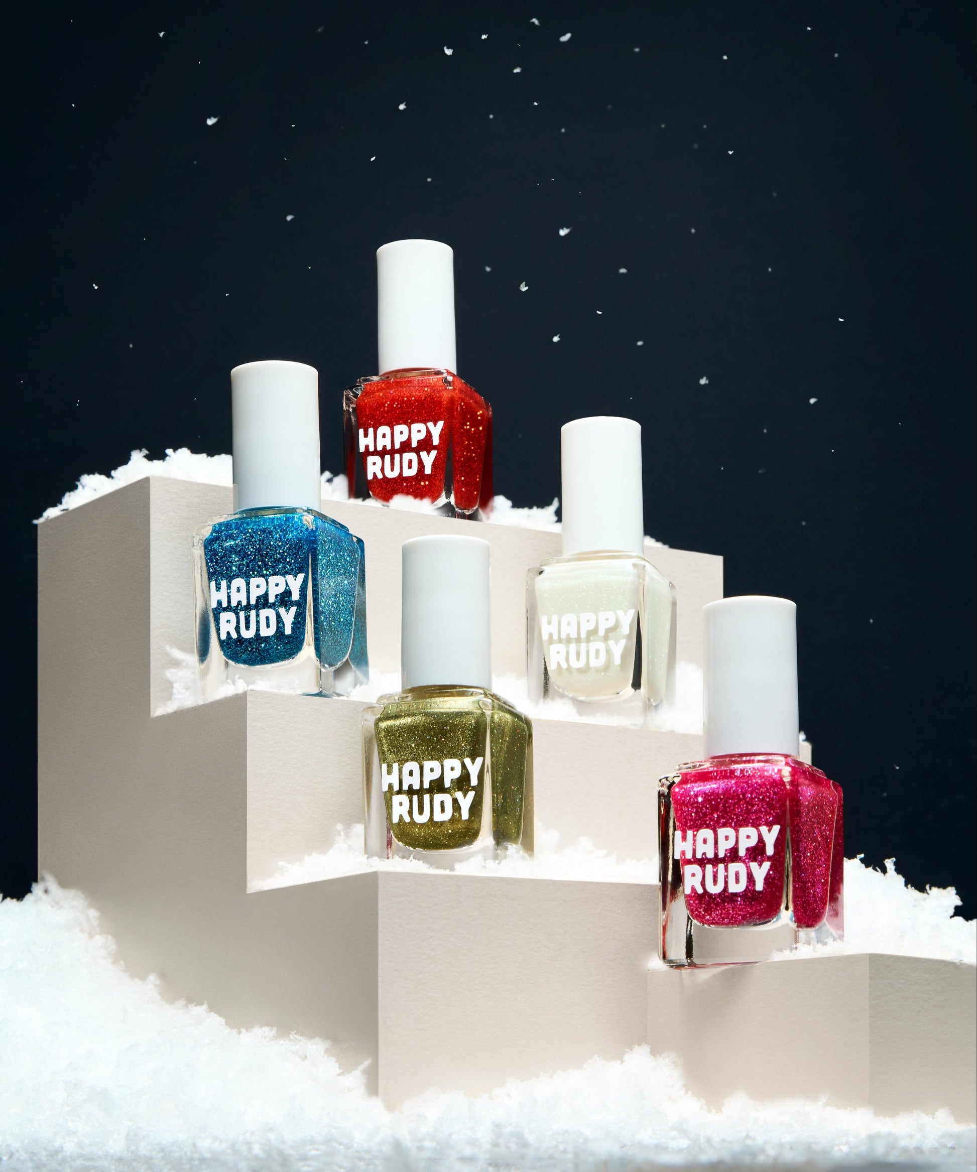 A collection of five nail polish bottles with glittery finishes displayed on a snowy surface, each with a different color glitter effect. The bottles are labeled with names such as 'Cerulean, Actually', 'Holiday Rebel', 'It's a Snow-derful Life', 'Keep the Change.