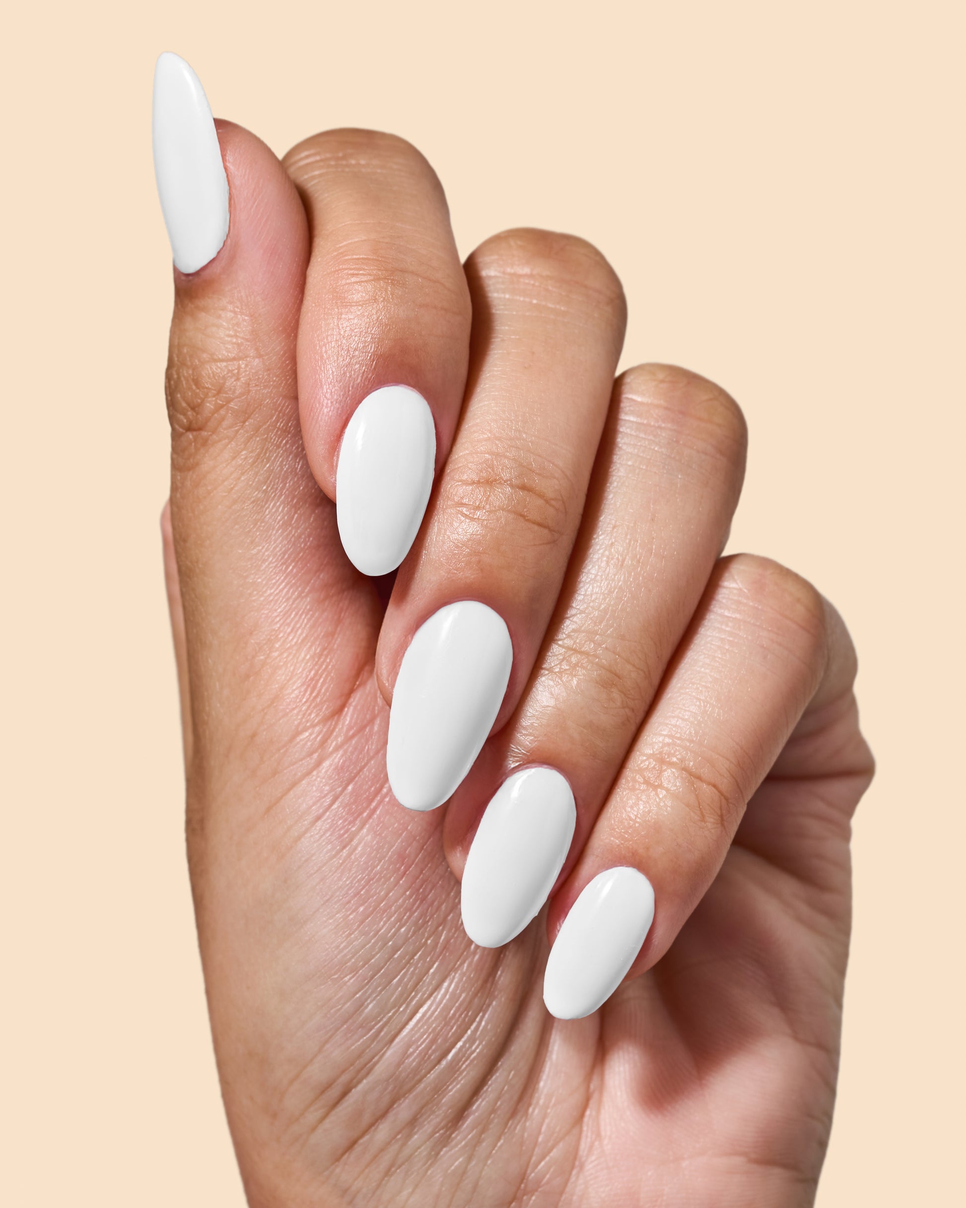 A close-up of a hand with long, almond-shaped nails painted in a glossy white manicure, set against a solid light beige background.