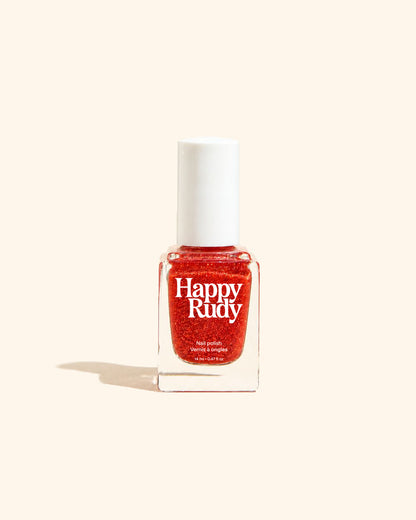 A bottle of Happy Rudy glitter nail polish in a vibrant red shade, set against a solid cream-colored background with a soft shadow to the left.