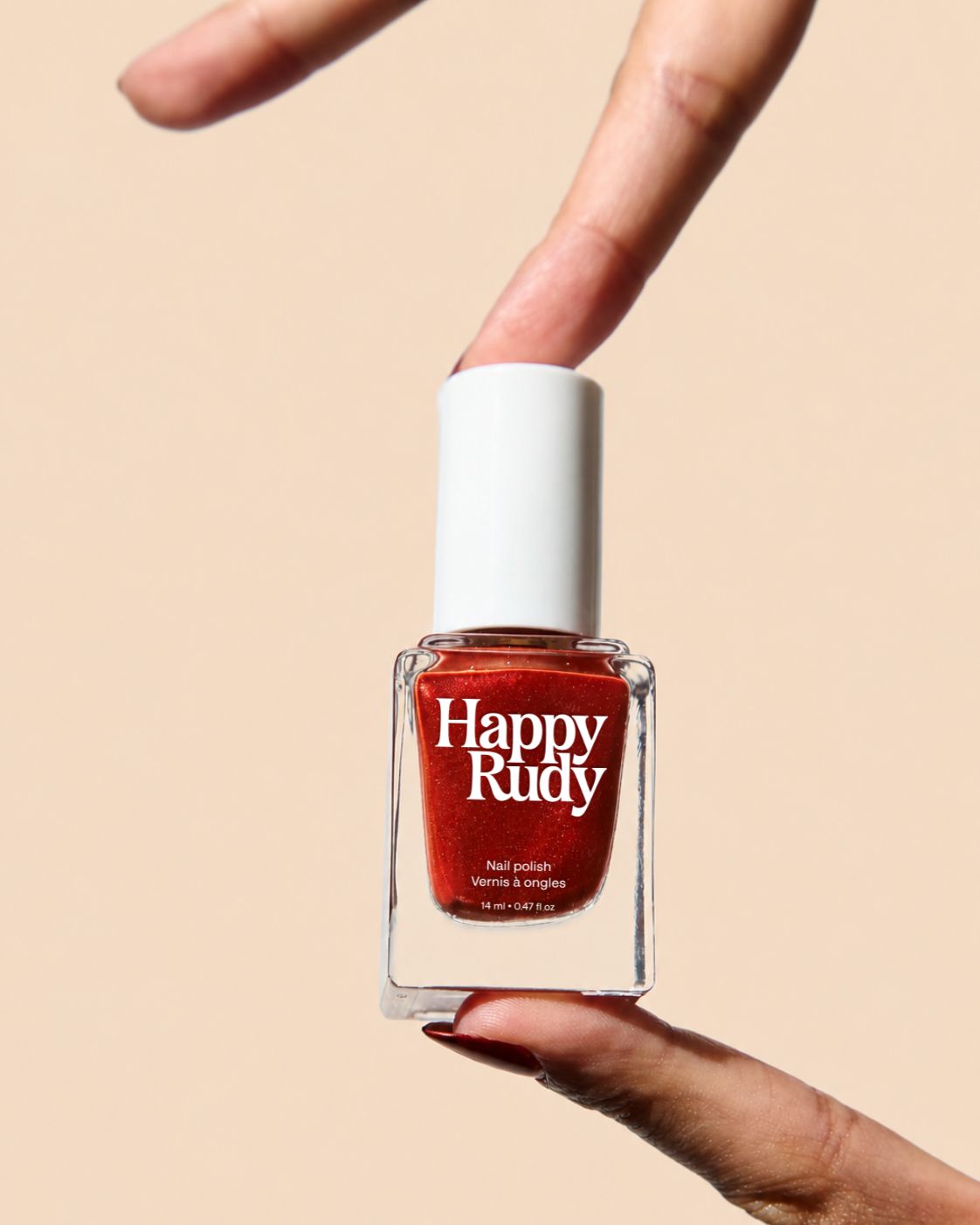 A bottle of Happy Rudy nail polish in a warm rust red shade, held between two hands against a solid beige background.