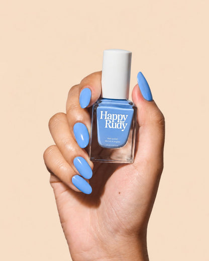 A hand with long, glossy light blue manicured nails holds a bottle of Happy Rudy nail polish against a solid beige background.