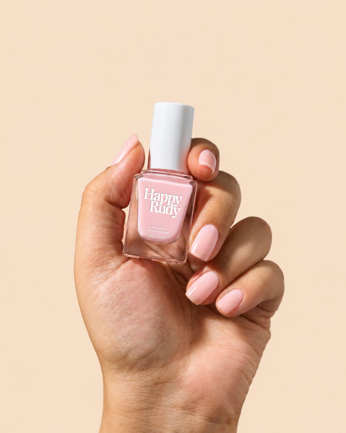 A hand holding a bottle of Happy Rudy nail polish in a light pink shade, set against a solid beige background.